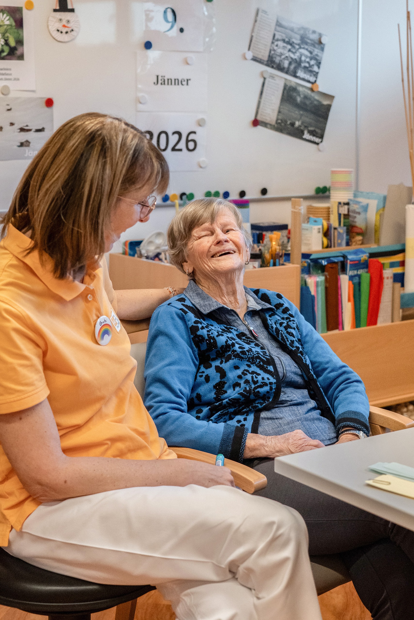 Mitarbeiterin sitzt neben einer Bewohnerin im Pflegeheim und spricht mit ihr.