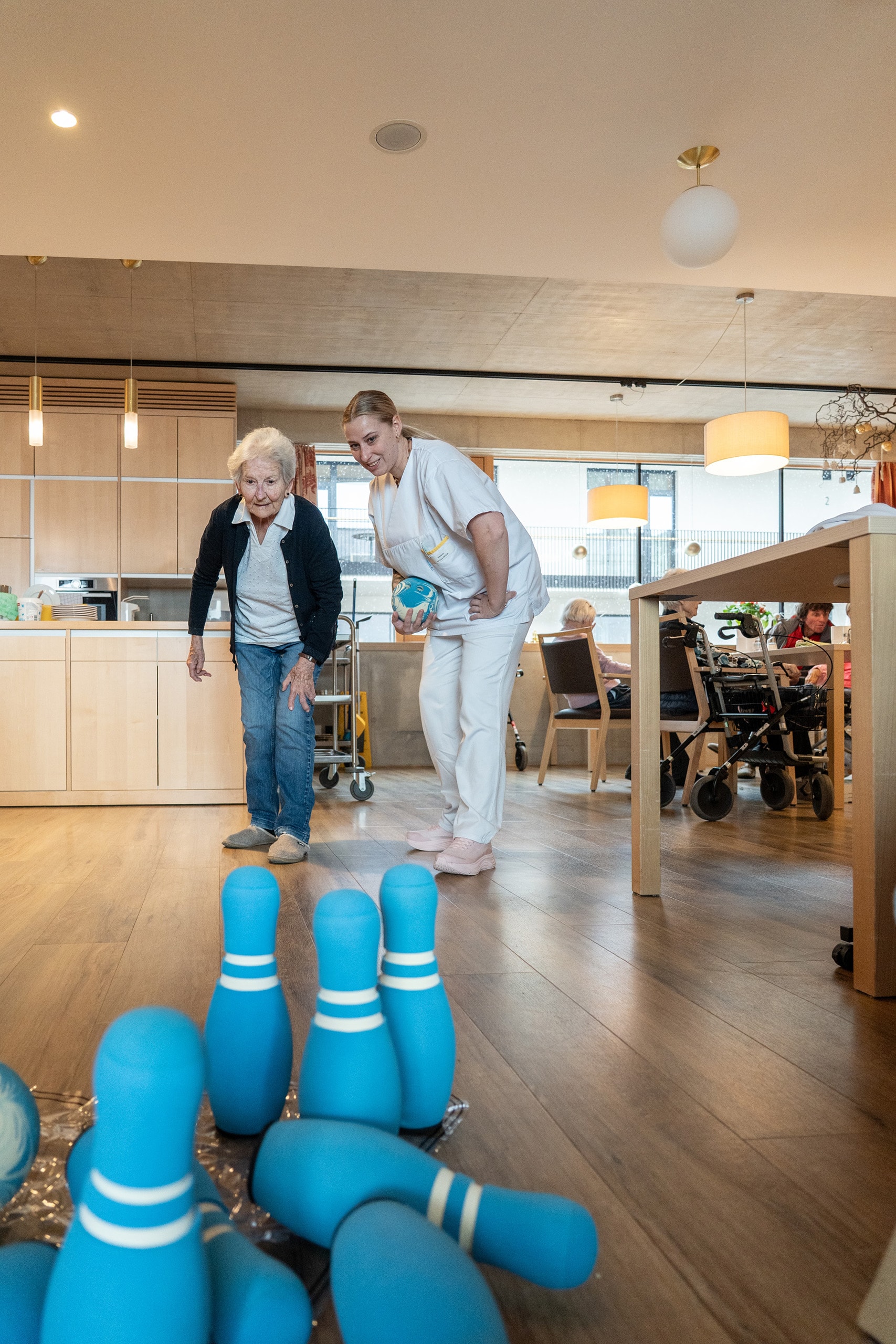 Bewohnerin spielt gemeinsam mit einer Betreuungsperson Kegeln im Aufenthaltsbereich eines Pflegeheims.