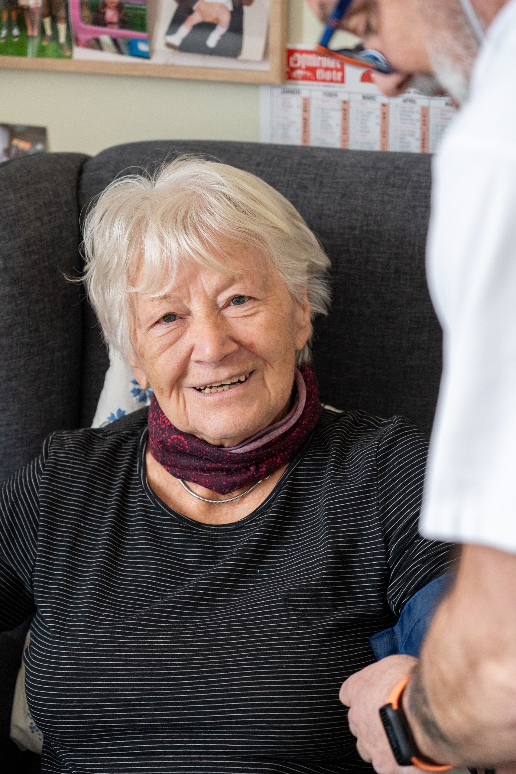 Bewohnerin sitzt in einem Sessel und lächelt eine Pflegekraft an, die neben ihr steht und mit ihr spricht.