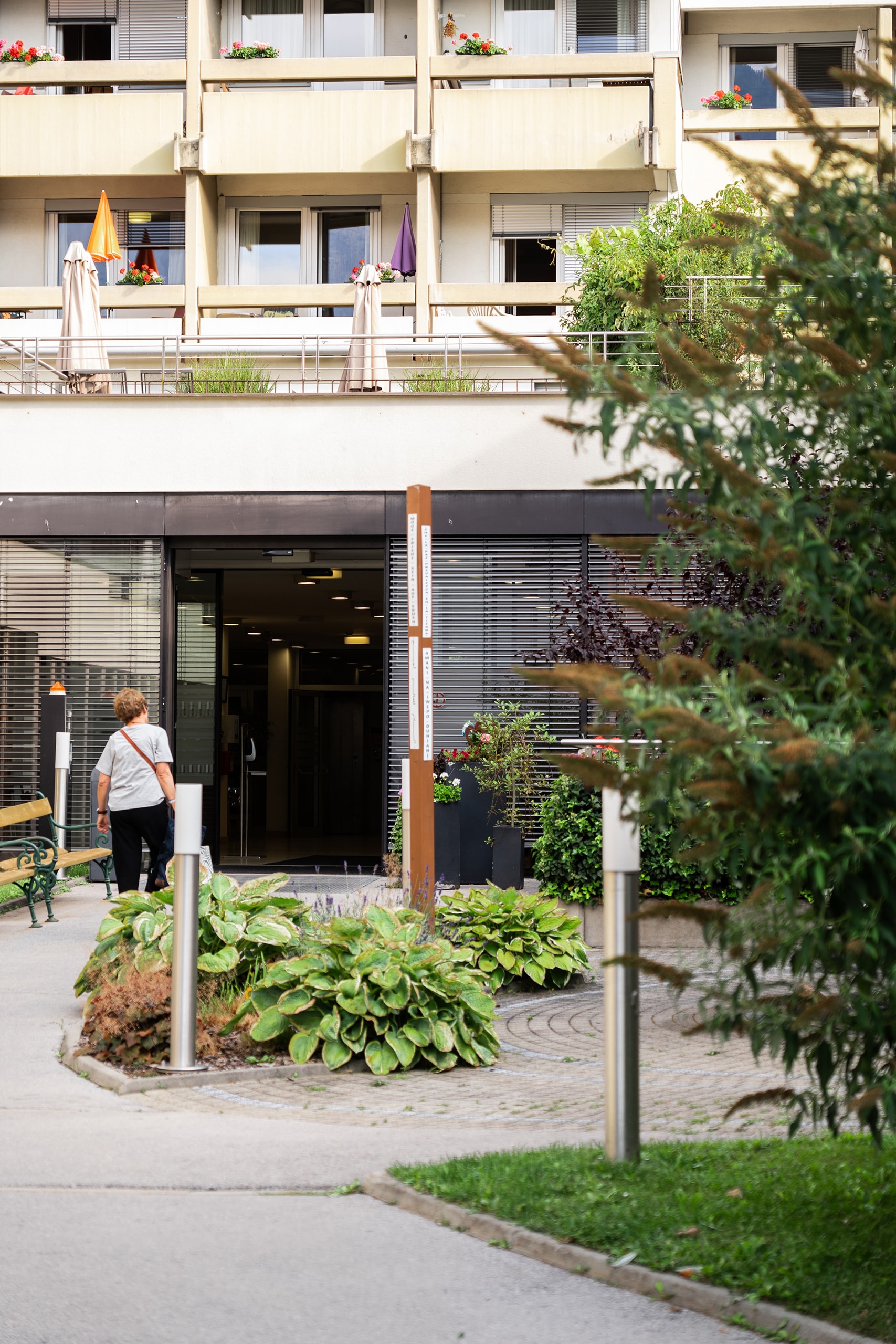 Eingangsbereich einer Wohn- und Pflegeeinrichtung (Wohn- & Pflegeheim Lienz) mit Gartenanlage; eine Person geht auf den Gebäudeeingang zu, Balkone mit Blumen im Hintergrund.