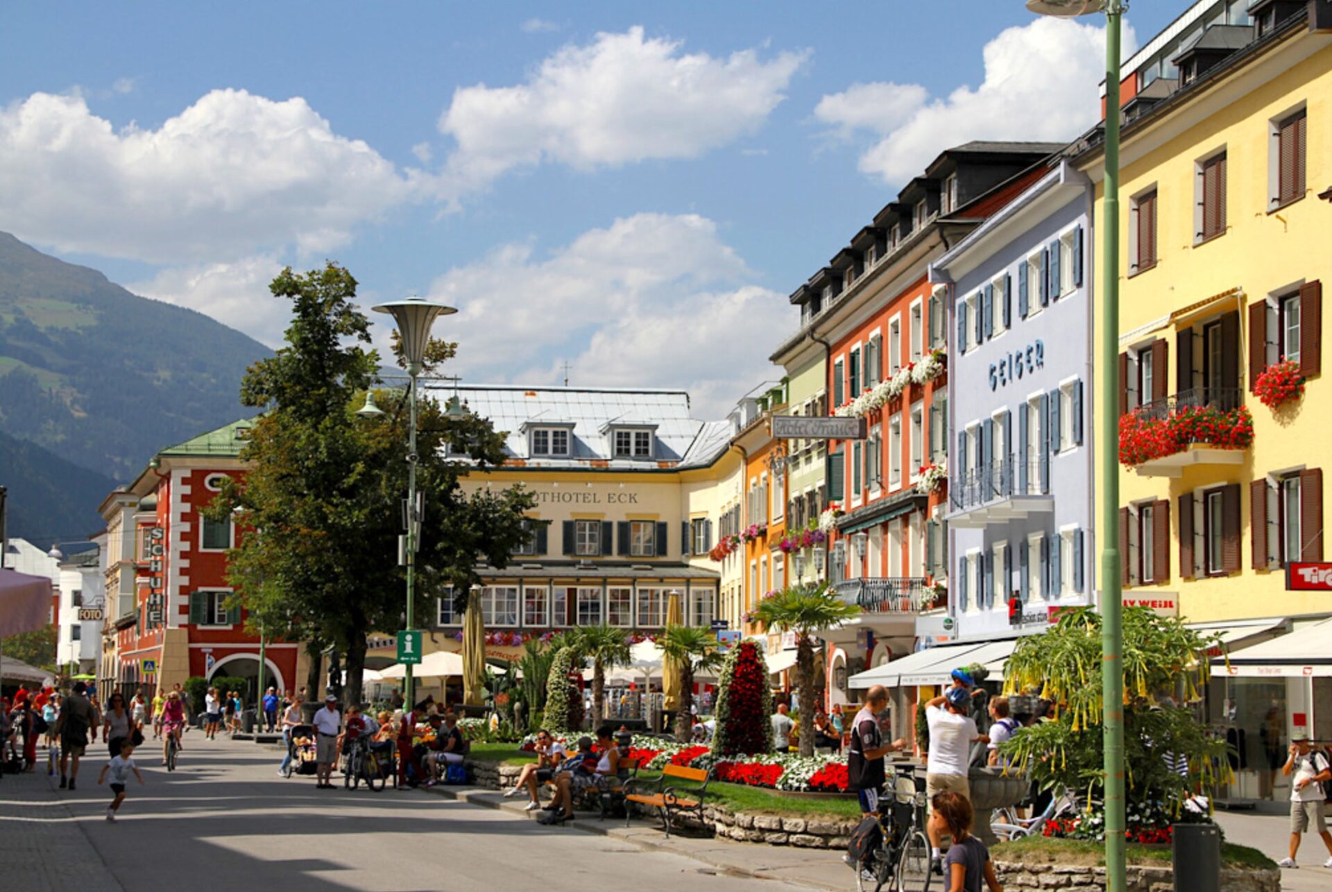 Belebter Stadtplatz mit bunten Häuserfassaden, Blumenbeeten, Sitzbänken und Menschen im Freien.