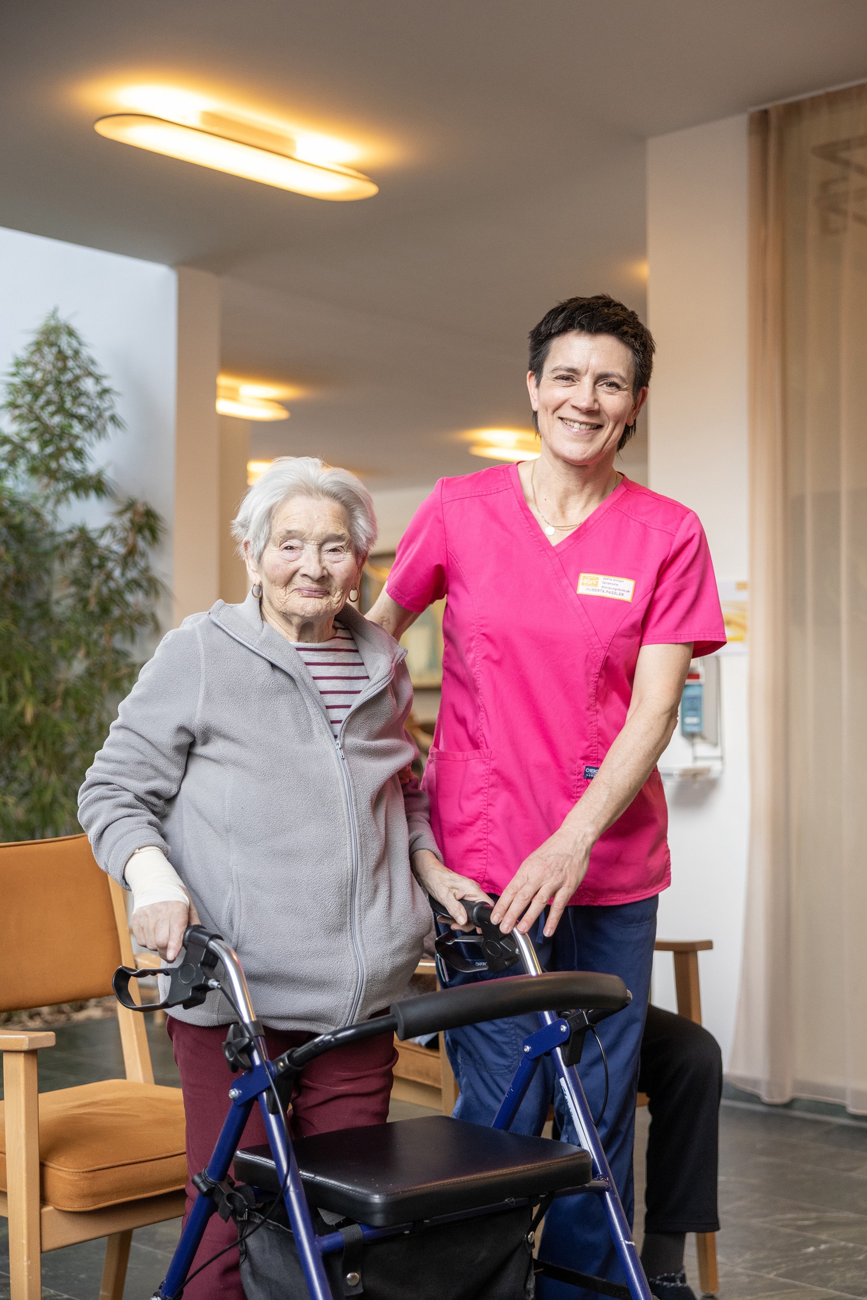 Pflegefachkraft begleitet eine ältere Bewohnerin mit Rollator im Wohn- und Pflegeheim, beide lächeln in die Kamera und stehen in einem hellen Gemeinschaftsbereich.