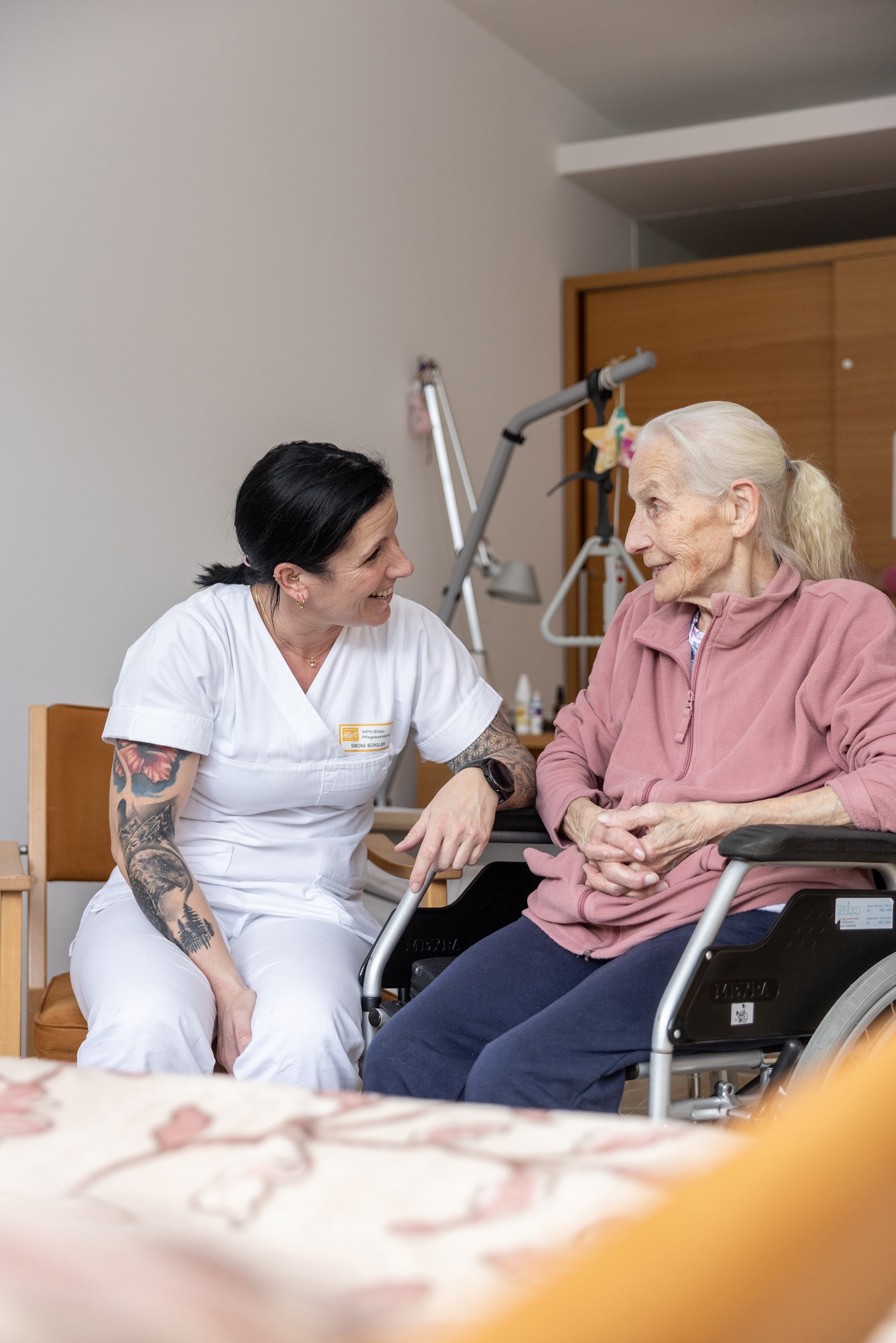 Pflegekraft sitzt neben einer Bewohnerin im Rollstuhl und führt ein Gespräch im Zimmer des Pflegeheims.