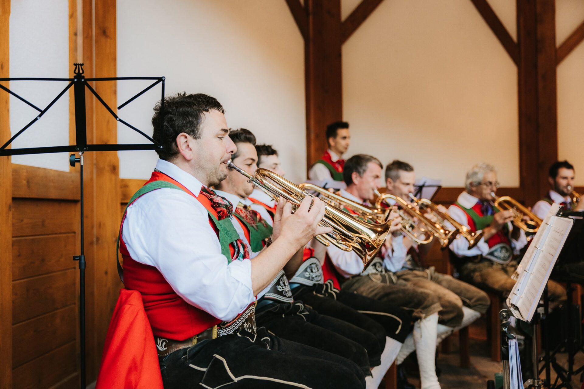 Mitglieder einer Musikkapelle in Tracht sitzen nebeneinander und spielen Trompeten bei einem Konzert in einem Holzsaal.