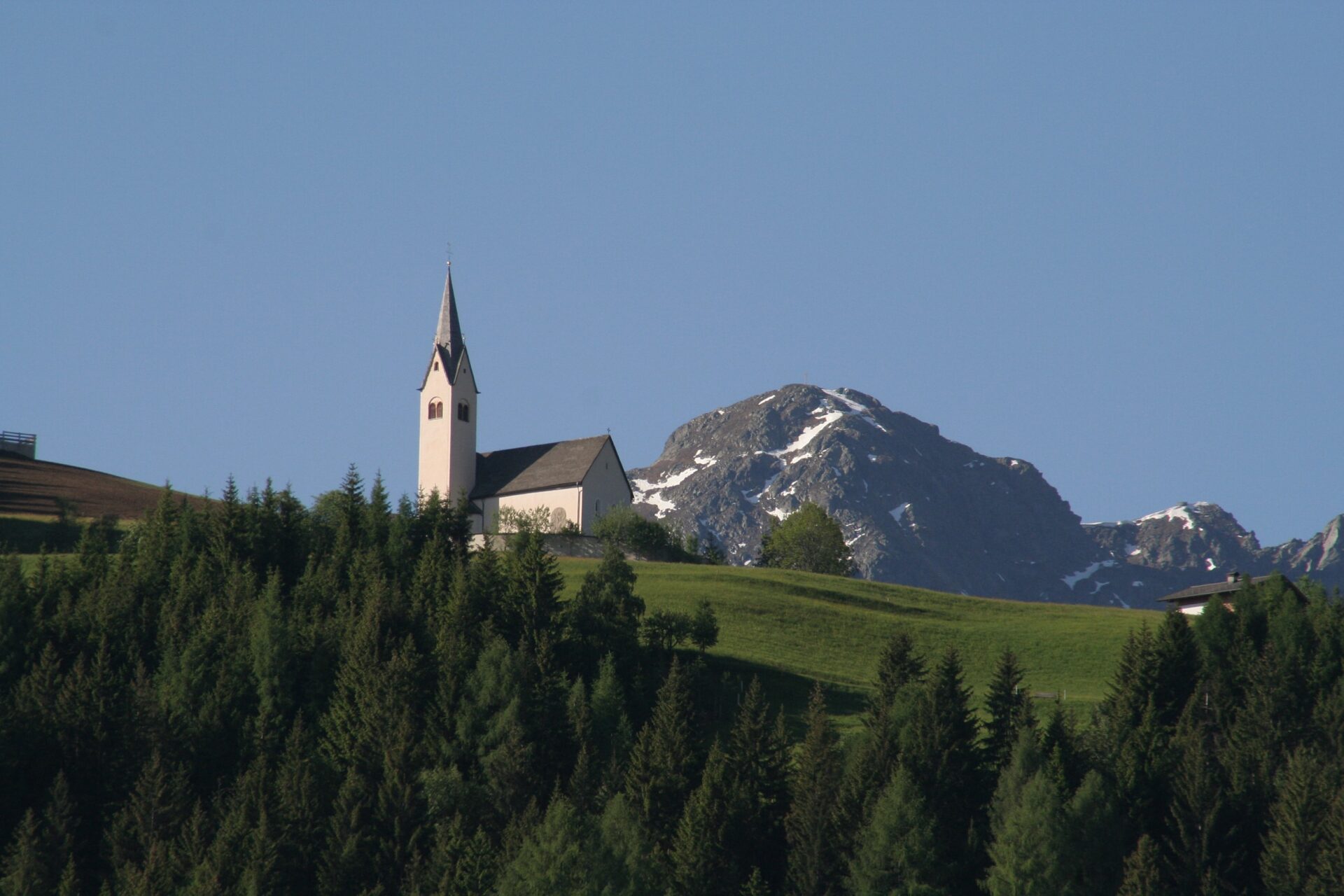 Kirche auf einem Hügel vor bewaldeter Landschaft und alpinem Bergpanorama.